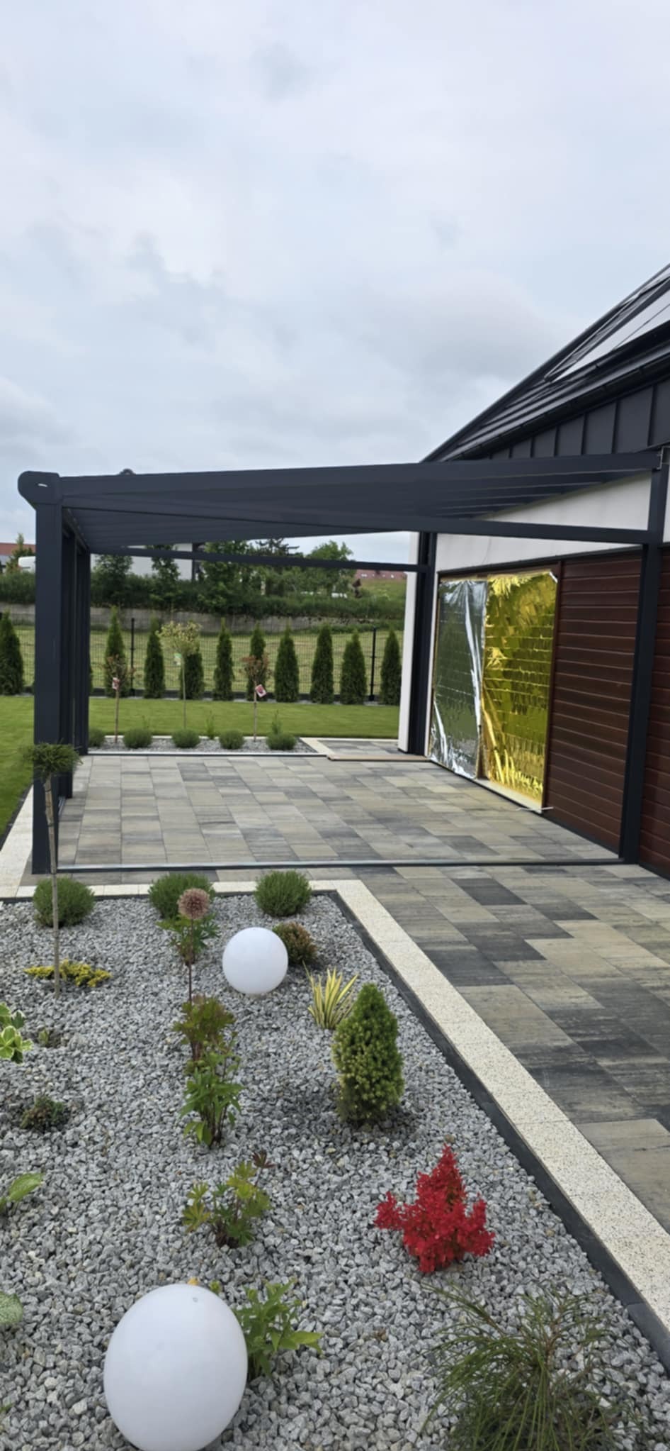 Nowoczesna pergola z kostki brukowej w szarościach, uzupełniona oświetleniem kulistym i rabatą z kamienia oraz roślin.  Widok na starannie utrzymany ogród.