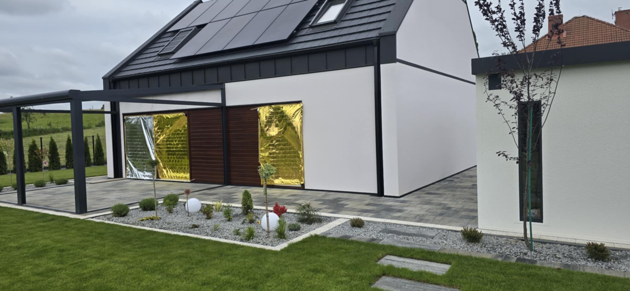 Nowoczesny dom z panelami solarnymi i zadaszeniem tarasu. Na posesji zadbany trawnik i kostka brukowa. Okna garażowe zabezpieczone folią.