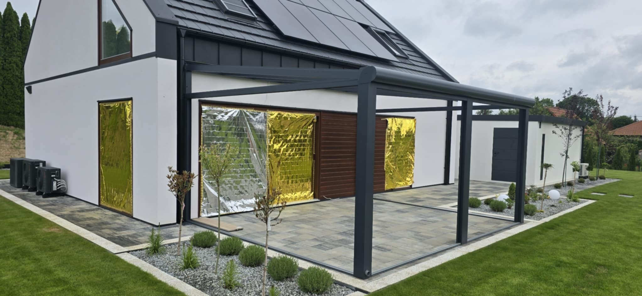 Nowoczesny dom z pergolą i kostką brukową. Okna zabezpieczone folią odblaskową. Zadbane trawniki i rabaty z kamienia ozdobnego. Panele słoneczne na dachu.