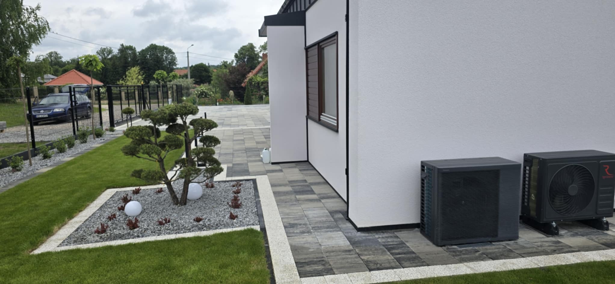 Nowoczesny ogród z kostką brukową w odcieniach szarości, ozdobnym drzewkiem bonsai w żwirowym obramowaniu i dwoma jednostkami zewnętrznymi klimatyzacji przy białej ścianie budynku.