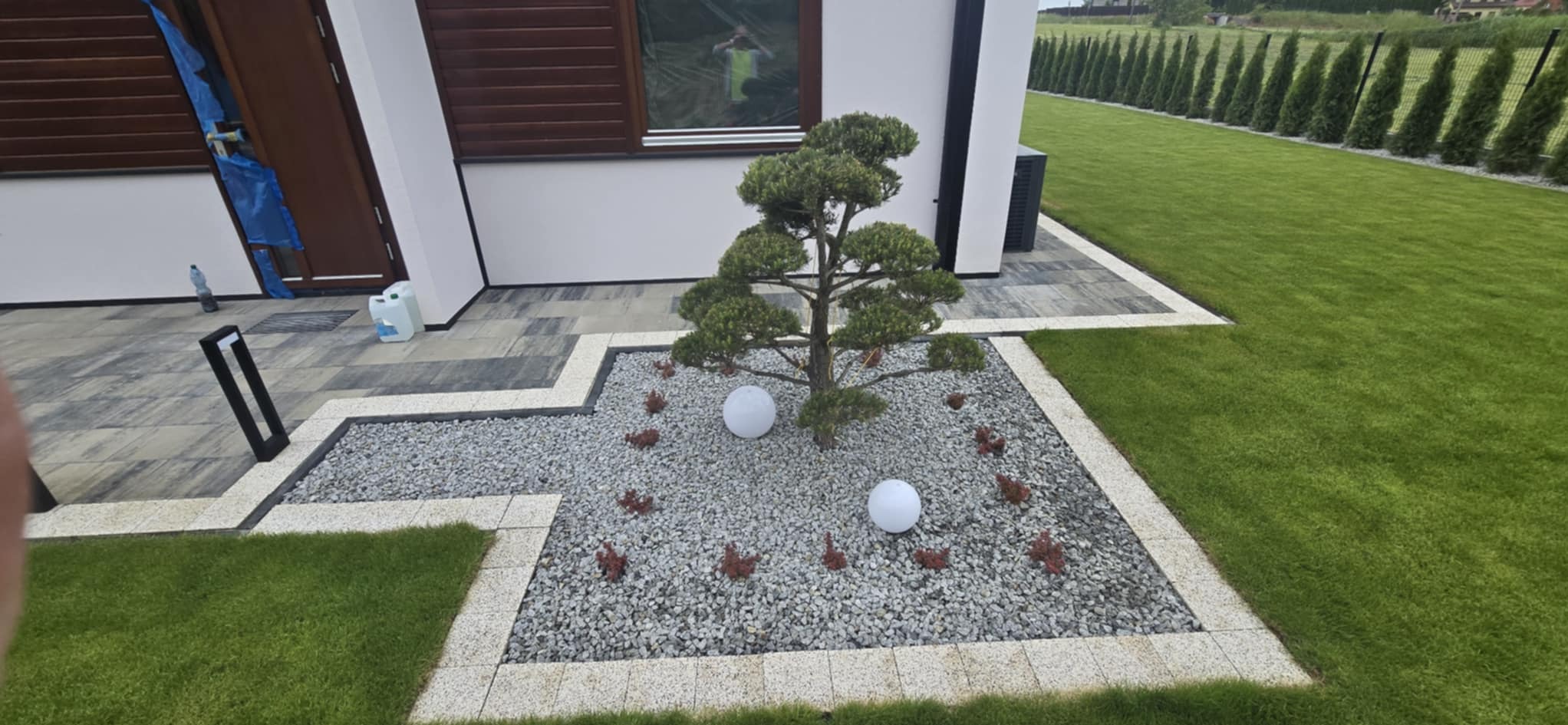 Ogród z ozdobnym drzewem bonsai, białymi kulami i szarym żwirem, otoczony kostką. Widoczny trawnik i rząd tuj w tle. Nowoczesny design ogrodu.