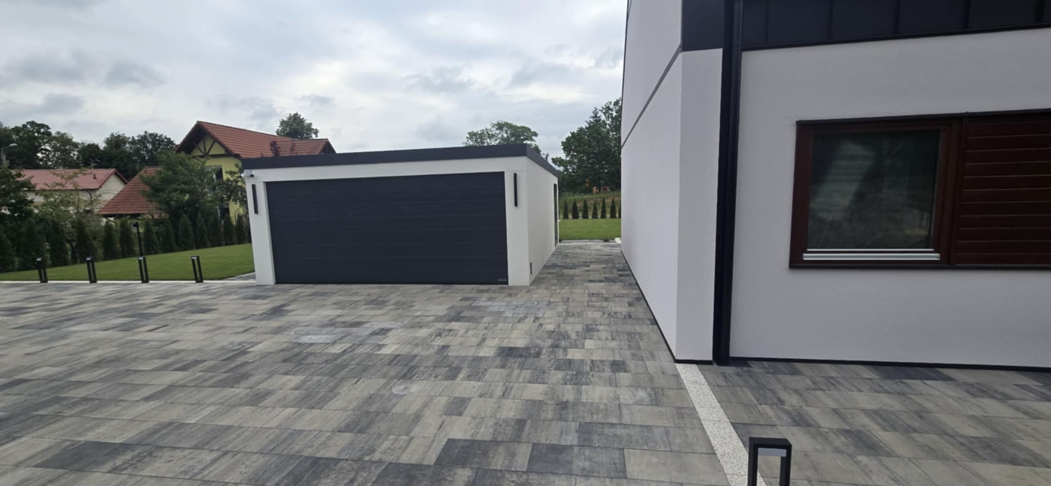 Ułożona kostka brukowa w szarościach prowadząca do garażu obok nowoczesnego domu z brązowymi oknami, w tle zieleń drzew i zabudowa jednorodzinna.