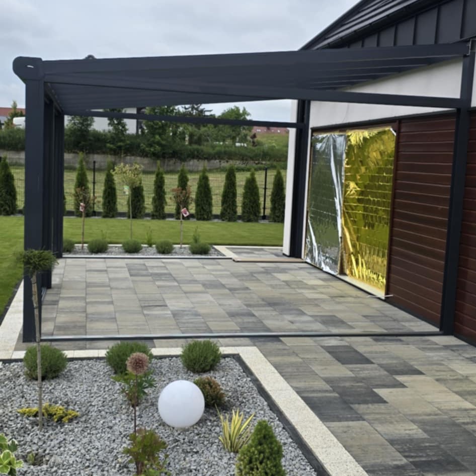 Nowoczesna pergola z szarą konstrukcją i kostką brukową w geometryczny wzór. Ogród z tujami i dekoracyjnym oświetleniem.  Zabezpieczone drzwi folią.