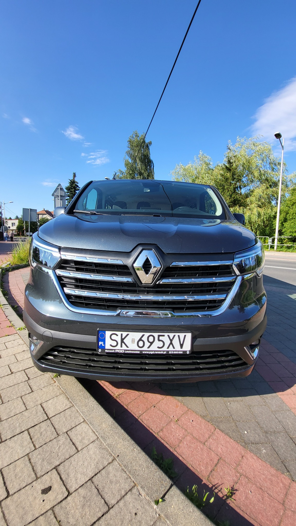 Przód ciemnoszarego Renault Espace zaparkowanego na chodniku z czerwonej kostki, widok na tablicę rejestracyjną SK 695XV, w tle błękitne niebo i zieleń drzew.