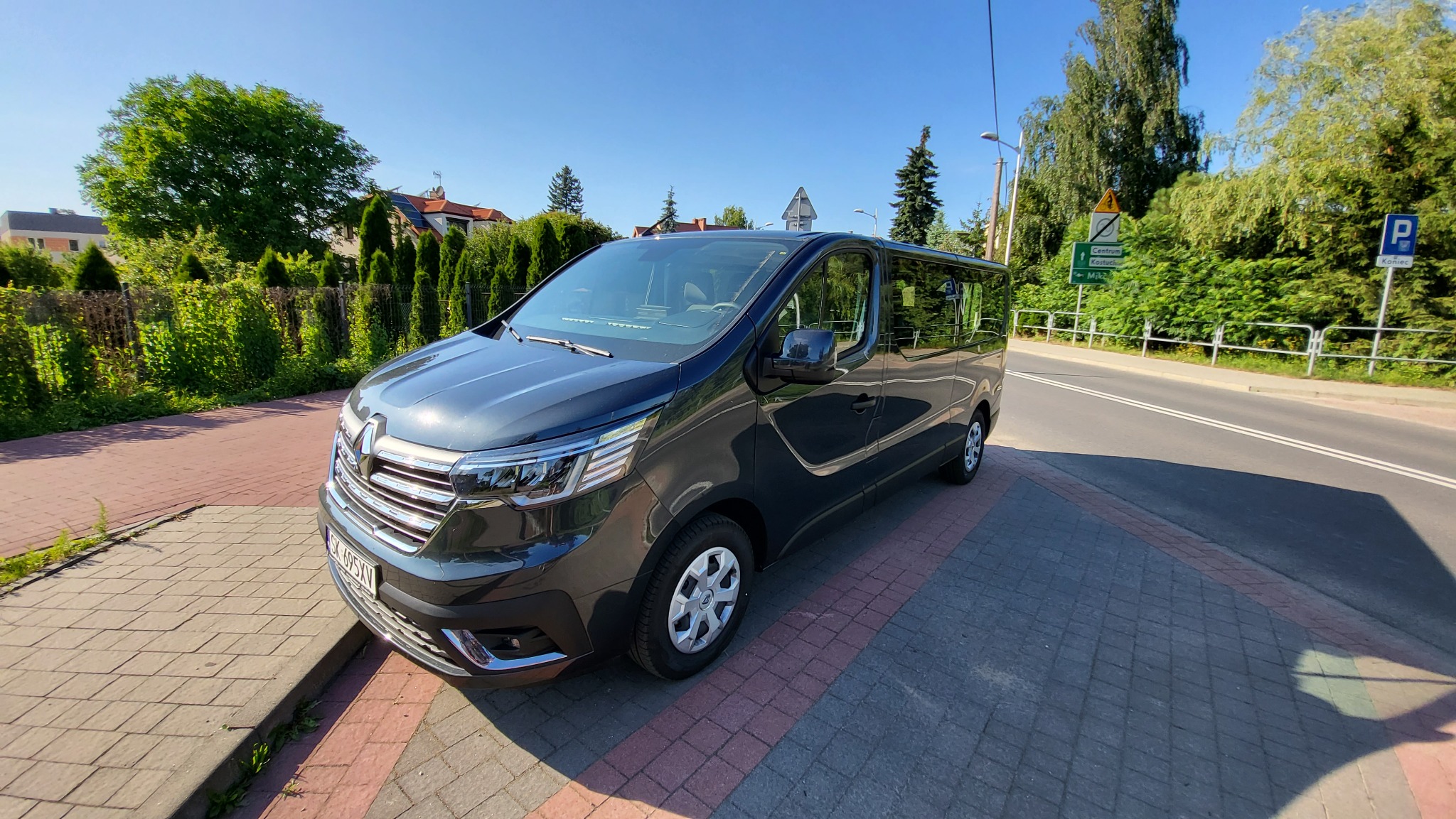 Ciemnoszary van Renault Trafic zaparkowany na chodniku z kostki, widok z przodu i boku, w tle ulica i zieleń.