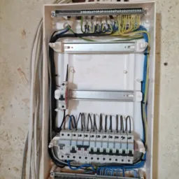 Otwarta skrzynka rozdzielcza z podłączonymi przewodami elektrycznymi na surowej ścianie, widoczne zabezpieczenia i listwy zaciskowe, kable opisane markerem.