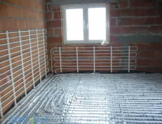 Rozłożone rury systemu ogrzewania podłogowego na folii izolacyjnej, przed wylewką. Widoczne mocowania do ściany z cegły i okno w tle.