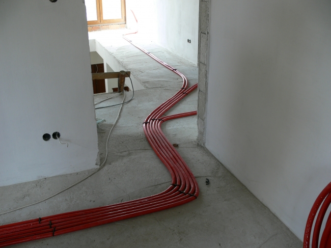 Czerwone rury instalacji grzewczej ułożone na surowym betonowym podłożu w trakcie budowy, widoczne otwory w ścianach na instalacje elektryczne.