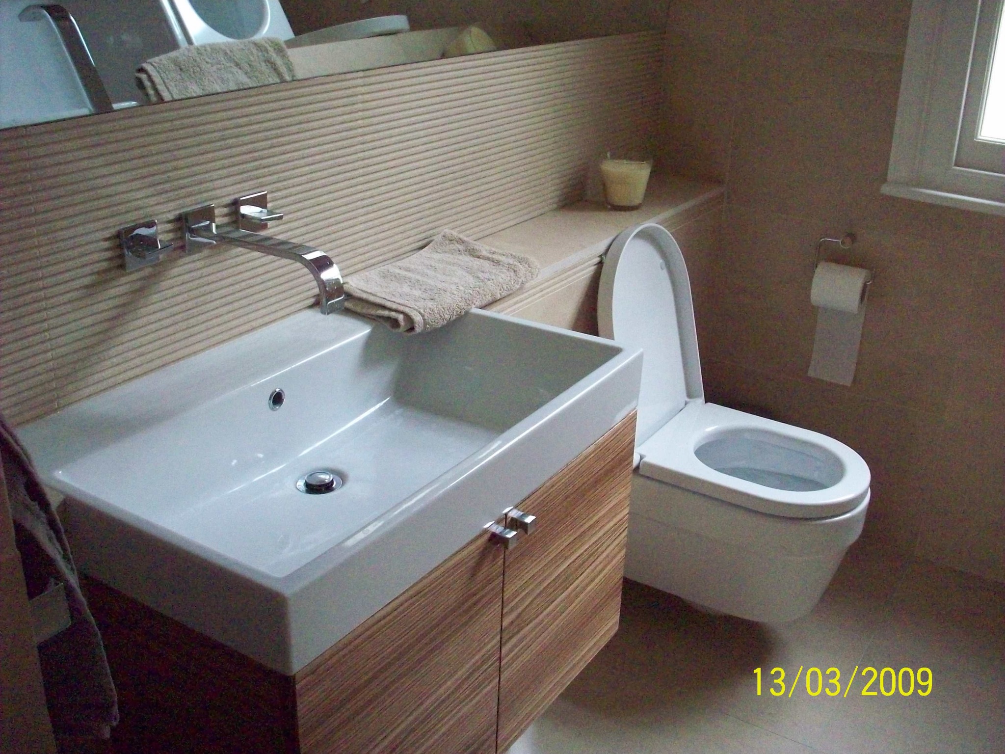 Nowoczesna łazienka z podwieszaną toaletą, umywalką nablatową na szafce z drewnopodobnym fornirem i chromowaną armaturą, wykończona w beżowych odcieniach, z widoczną datą na podłodze.