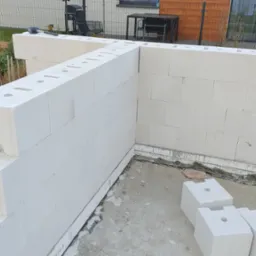 Wznoszenie muru fundamentowego z białych bloczków betonowych na fundamencie, widoczne narzędzia i fragment ogrodu w tle.