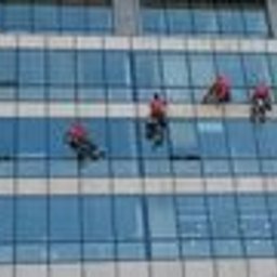 Prace wysokościowe - profesjonalne usługi alpinistyczne - Pracownicy alpinistyczni w różowych kombinezonach myją szklaną fasadę wysokiego biurowca, widoczne odbicia chmur w oknach.