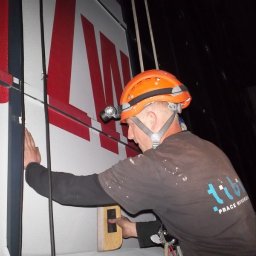 Prace wysokościowe - profesjonalne usługi alpinistyczne - Pracownik w pomarańczowym kasku z czołówką, zabezpieczony linami, dokonuje pomiarów na elewacji budynku z dużym czerwonym napisem, używając poziomicy.