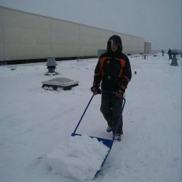 Prace wysokościowe - profesjonalne usługi alpinistyczne - Osoba w kurtce odśnieża płaski dach budynku przemysłowego przy użyciu niebieskiego odśnieżacza na kółkach. Na dachu widoczne świetliki i wentylatory, a w tle zarys innych osób.