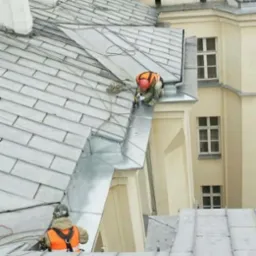 Pracownicy w kamizelkach odblaskowych zabezpieczają rynny na dachu kamienicy z jasną elewacją i oknami.