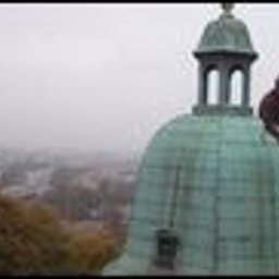 Prace wysokościowe - profesjonalne usługi alpinistyczne - Pracownik w uprzęży, siedzący na miedzianej kopule wieży kościoła podczas prac konserwatorskich, z widocznymi śladami patyny i farbą w wiaderku. W tle zamglony krajobraz miejski.