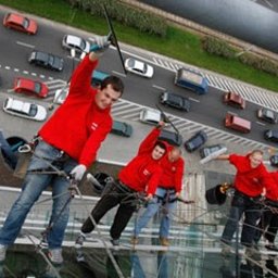 Prace wysokościowe - profesjonalne usługi alpinistyczne - Grupa mężczyzn w czerwonych koszulkach i jeansach, zabezpieczona linami, czyści szklaną elewację budynku z widokiem na ruchliwą ulicę z samochodami w tle.