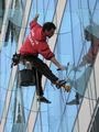 Pracownik w czerwonej bluzie, zabezpieczony linami, czyści szklaną elewację wysokiego budynku za pomocą mopa na kiju i wiadra z wodą.