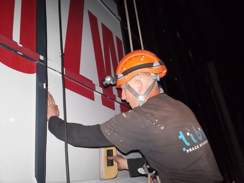 Pracownik w pomarańczowym kasku z czołówką, zabezpieczony linami, dokonuje pomiarów na elewacji budynku z dużym czerwonym napisem, używając poziomicy.