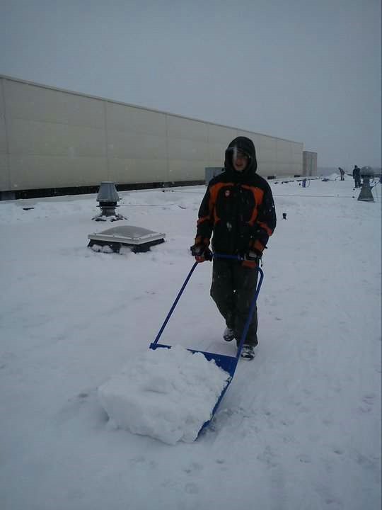 Osoba w kurtce odśnieża płaski dach budynku przemysłowego przy użyciu niebieskiego odśnieżacza na kółkach. Na dachu widoczne świetliki i wentylatory, a w tle zarys innych osób.