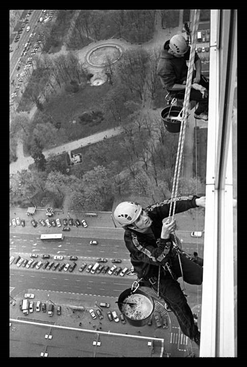 Czarno-białe zdjęcie dwóch alpinistów przemysłowych na wysokościach, myjących okna wieżowca z widokiem na park z fontanną i ruchliwą ulicę z zaparkowanymi samochodami.