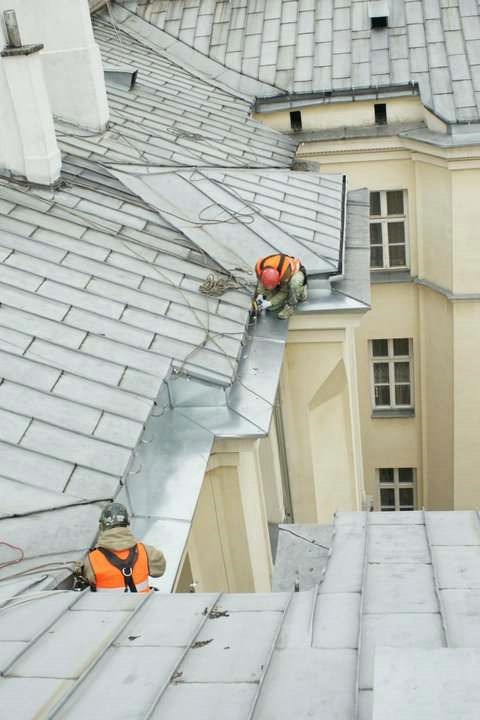 Pracownicy w kamizelkach odblaskowych zabezpieczają rynny na dachu kamienicy z jasną elewacją i oknami.