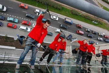 Grupa mężczyzn w czerwonych koszulkach i jeansach, zabezpieczona linami, czyści szklaną elewację budynku z widokiem na ruchliwą ulicę z samochodami w tle.