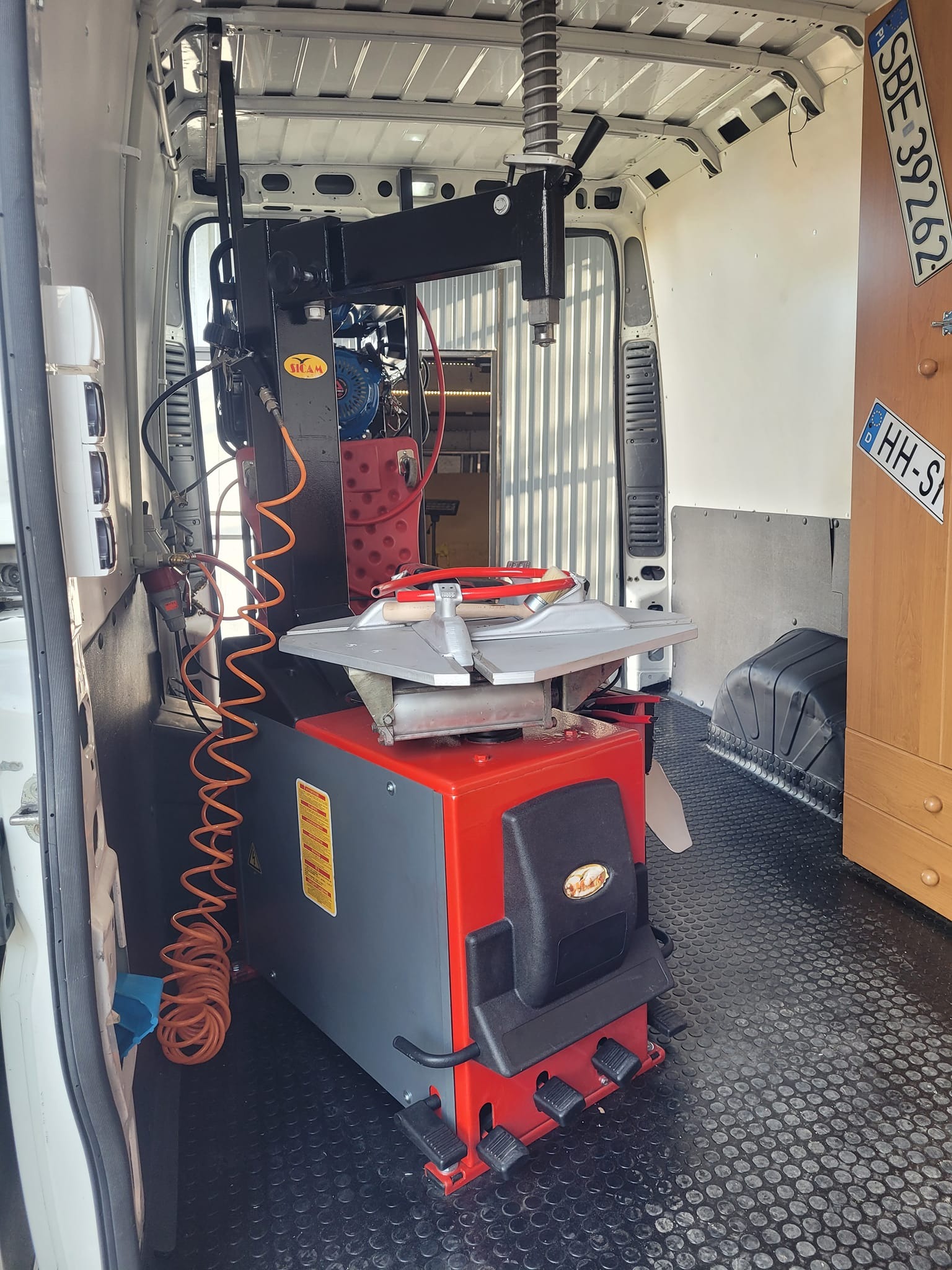 Mobilny warsztat wewnątrz furgonetki: montażownica do opon z pomarańczowym przewodem pneumatycznym, tablice rejestracyjne na szafce z tyłu.