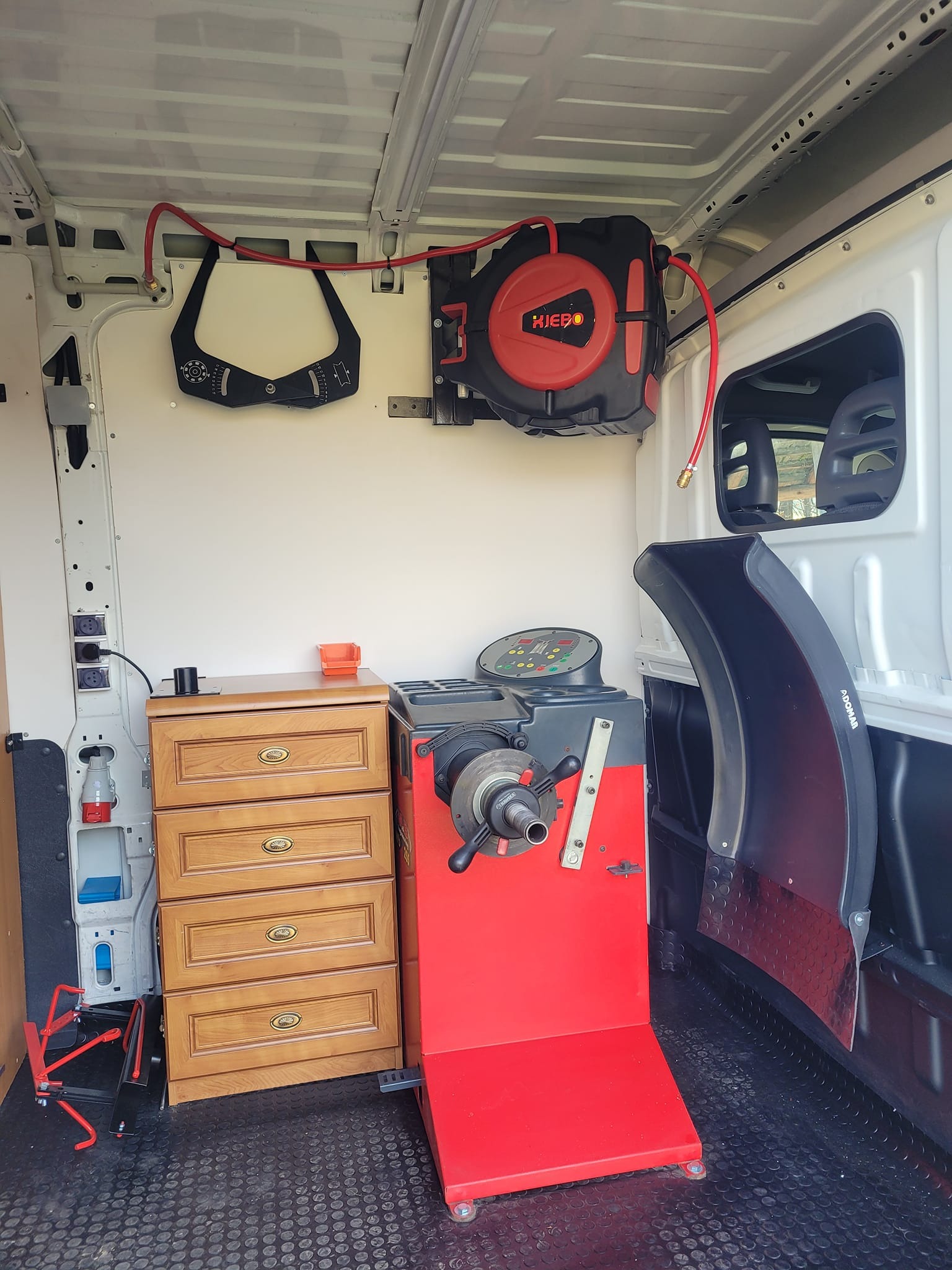 Mobilny warsztat wewnątrz furgonetki: wyważarka do kół, szafka narzędziowa z drewnianymi szufladami, zwijak do węża pneumatycznego HIEBO zamontowany na ścianie, czarny uchwyt do pomiaru szerokości...