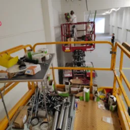 Pracownik w kombinezonie ochronnym i słuchawkach na podnośniku koszowym wierci otwory w suficie, widok z drugiego podnośnika z narzędziami i materiałami na platformie.