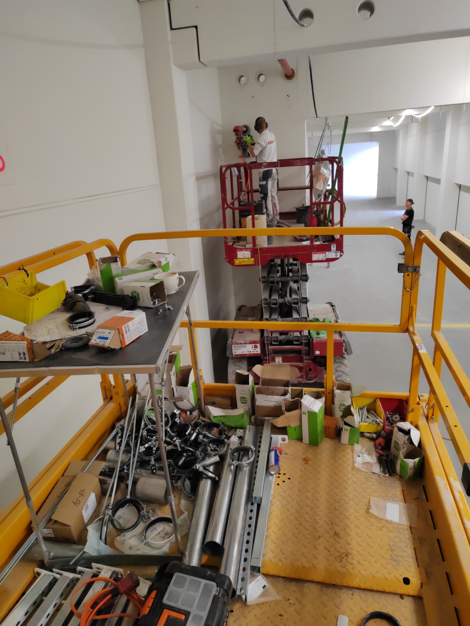 Pracownik w kombinezonie ochronnym i słuchawkach na podnośniku koszowym wierci otwory w suficie, widok z drugiego podnośnika z narzędziami i materiałami na platformie.