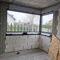 Narożne okno w surowym stanie, widok na ogród z tujami i domem z czerwonym dachem, ściany z betonu komórkowego.