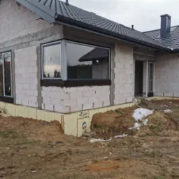 Surowy stan budynku z białego bloczka z zamontowanymi oknami w czarnych ramach, widoczna izolacja fundamentów i częściowo wyrównany teren.