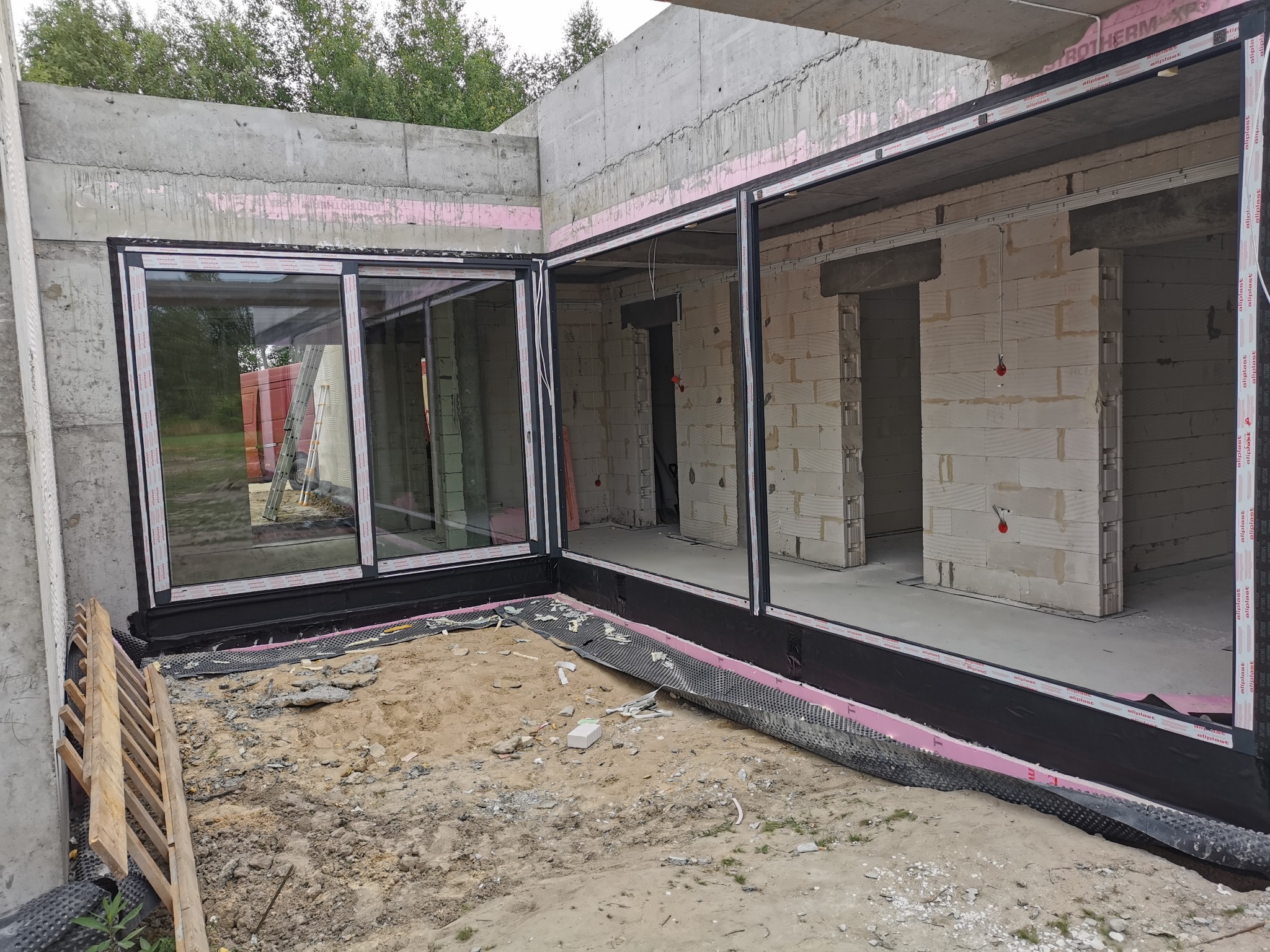 Montaż okien w surowym stanie budynku, widoczne detale konstrukcyjne i materiały izolacyjne, w tle drabina i kontener.