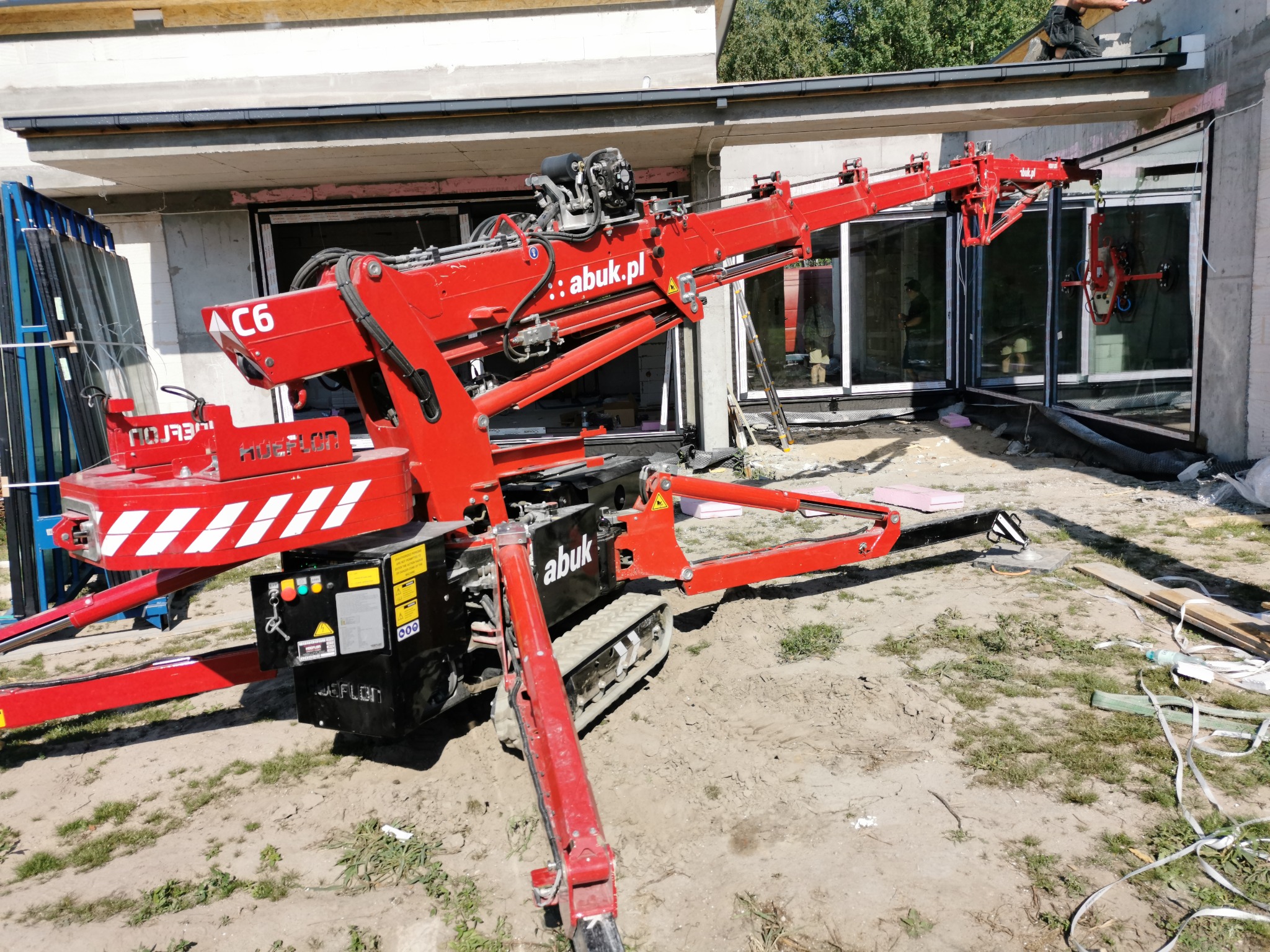 Montaż okien w Lublinie z użyciem czerwonego mini żurawia gąsienicowego z przyssawkami do szkła, widoczne surowe ściany budynku w trakcie budowy.