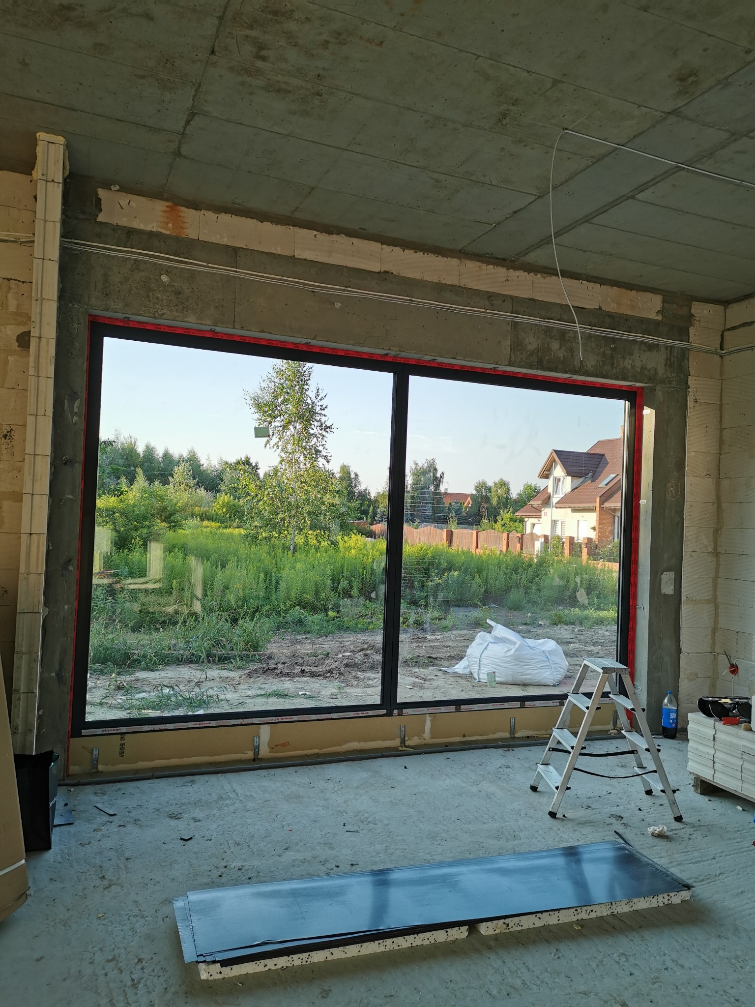 Montaż dużego okna aluminiowego z widokiem na ogród i dom, widoczna drabina i materiały budowlane w surowym wnętrzu.