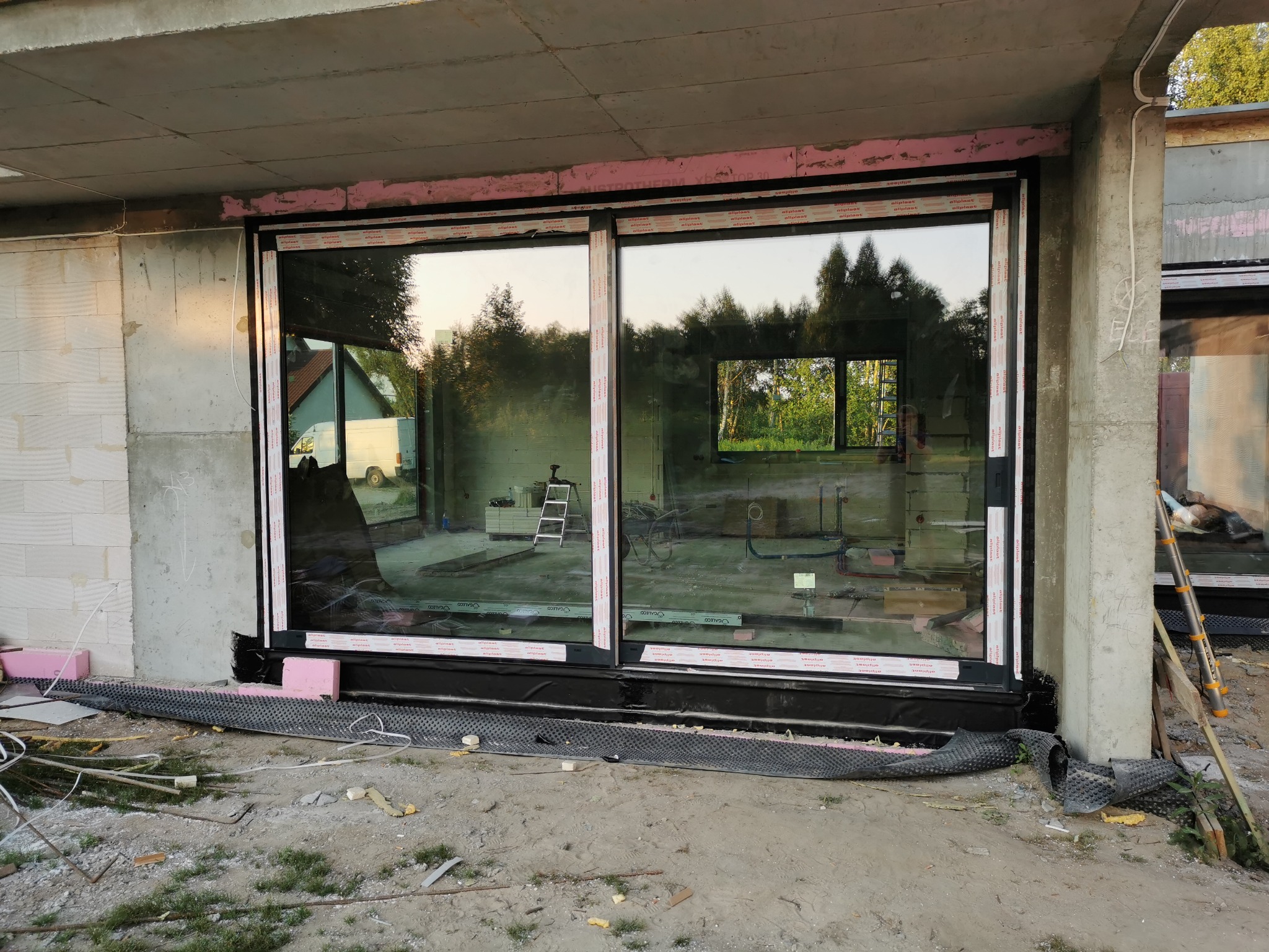 Duże przesuwne okno w czarnej ramie, zamontowane w surowym stanie budynku, widoczne materiały budowlane i drabina w tle.