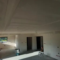 Wnętrze w trakcie prac wykończeniowych: ściany z widocznymi puszkami elektrycznymi, częściowo wykończony sufit podwieszany z płyt kartonowo-gipsowych, drabina i materiały budowlane na podłodze.