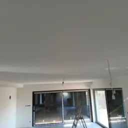 Wnętrze po nałożeniu gładzi, widoczne ściany i sufit w kolorze białym, drabina oparta o duże okno przesuwne z roletami zewnętrznymi, wystające kable oświetleniowe.