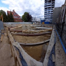 Obsługa Inwestycji Budowlanych Paweł Lemańczyk - Wykonanie wykopu pod budowę z zabezpieczeniem ścian wykopu rurami stalowymi. W tle budynki mieszkalne i biurowe. Widok z góry na plac budowy.