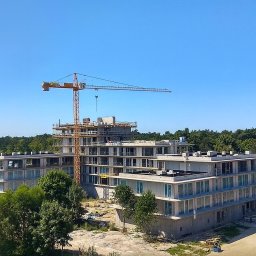 Obsługa Inwestycji Budowlanych Paweł Lemańczyk - Wysoki budynek w trakcie budowy, widoczny żuraw budowlany na tle błękitnego nieba i zieleni drzew. Budynek z licznymi oknami i balkonami w surowym stanie.