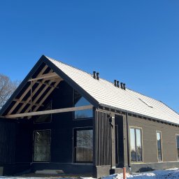 Obsługa Inwestycji Budowlanych Paweł Lemańczyk - Dom w trakcie budowy, z widoczną konstrukcją dachu i oknami. Elewacja pokryta czarną membraną, dach częściowo ośnieżony. Błękitne niebo w tle.