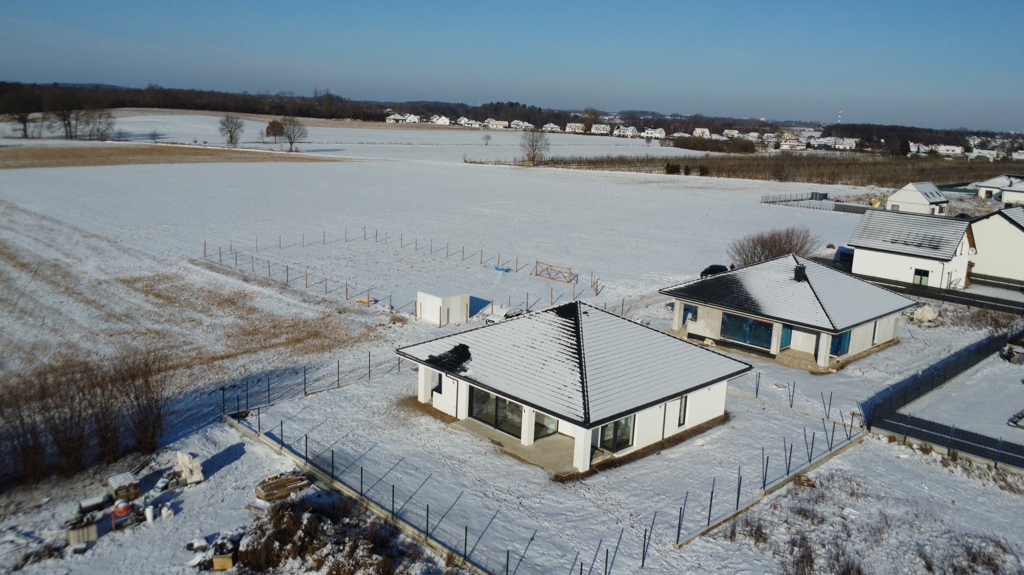 Zimowy krajobraz z nowoczesnym, parterowym domem z ciemną dachówką, otoczonym śniegiem i ogrodzeniem. Widok z lotu ptaka na osiedle w tle.