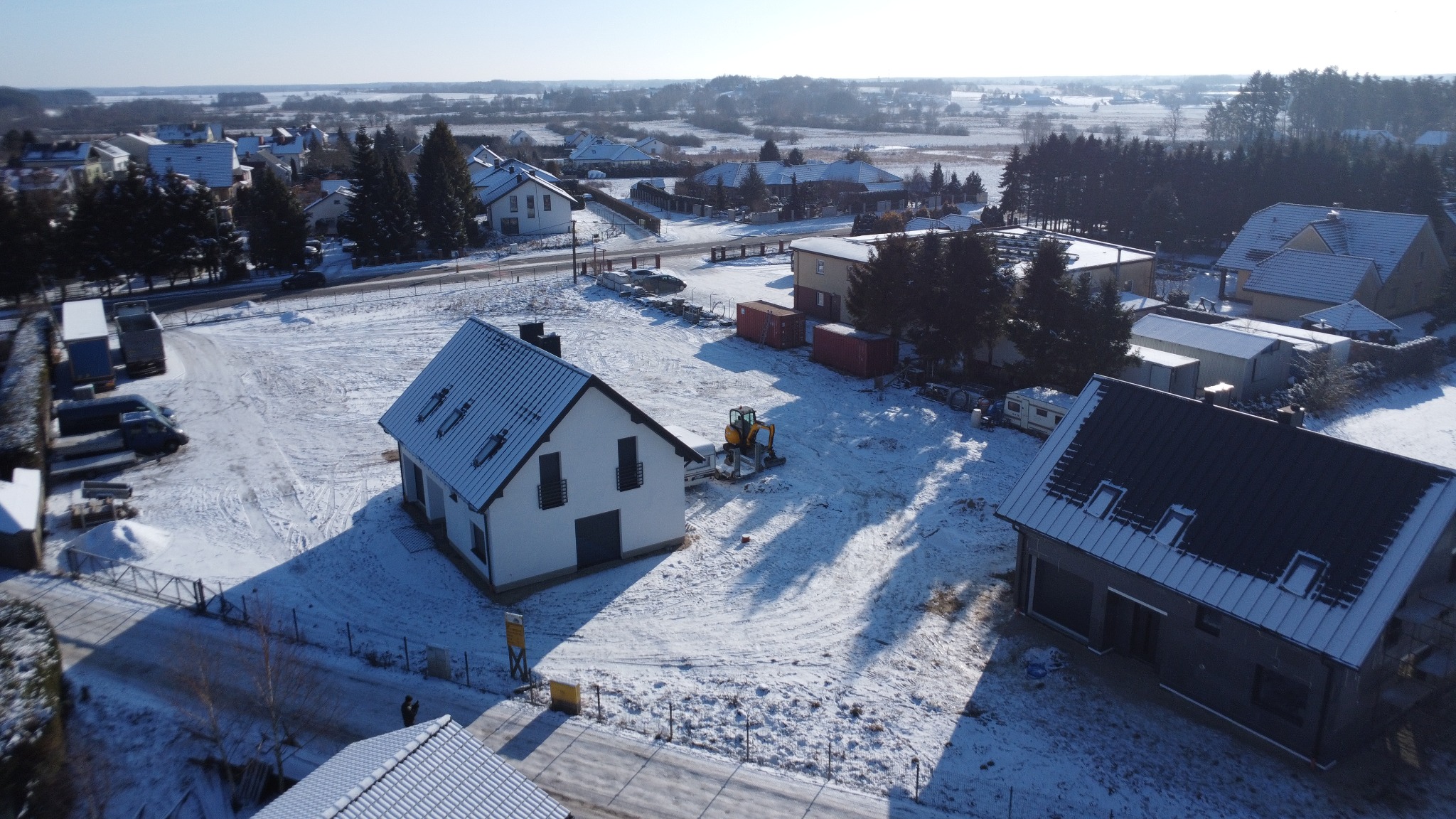 Zimowy krajobraz z nowo wybudowanym domem jednorodzinnym z ciemną stolarką okienną i dachem, widzianym z lotu ptaka. Na posesji stoi mała koparka.