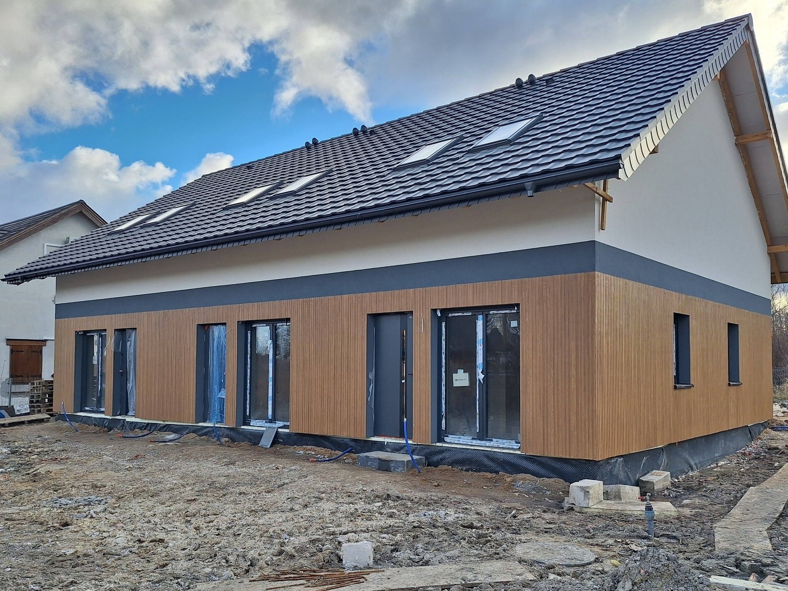 Nowoczesny dom jednorodzinny w trakcie wykończenia elewacji z drewnianą okładziną, antracytową stolarką i dachem z lukarnami, na tle pochmurnego nieba.