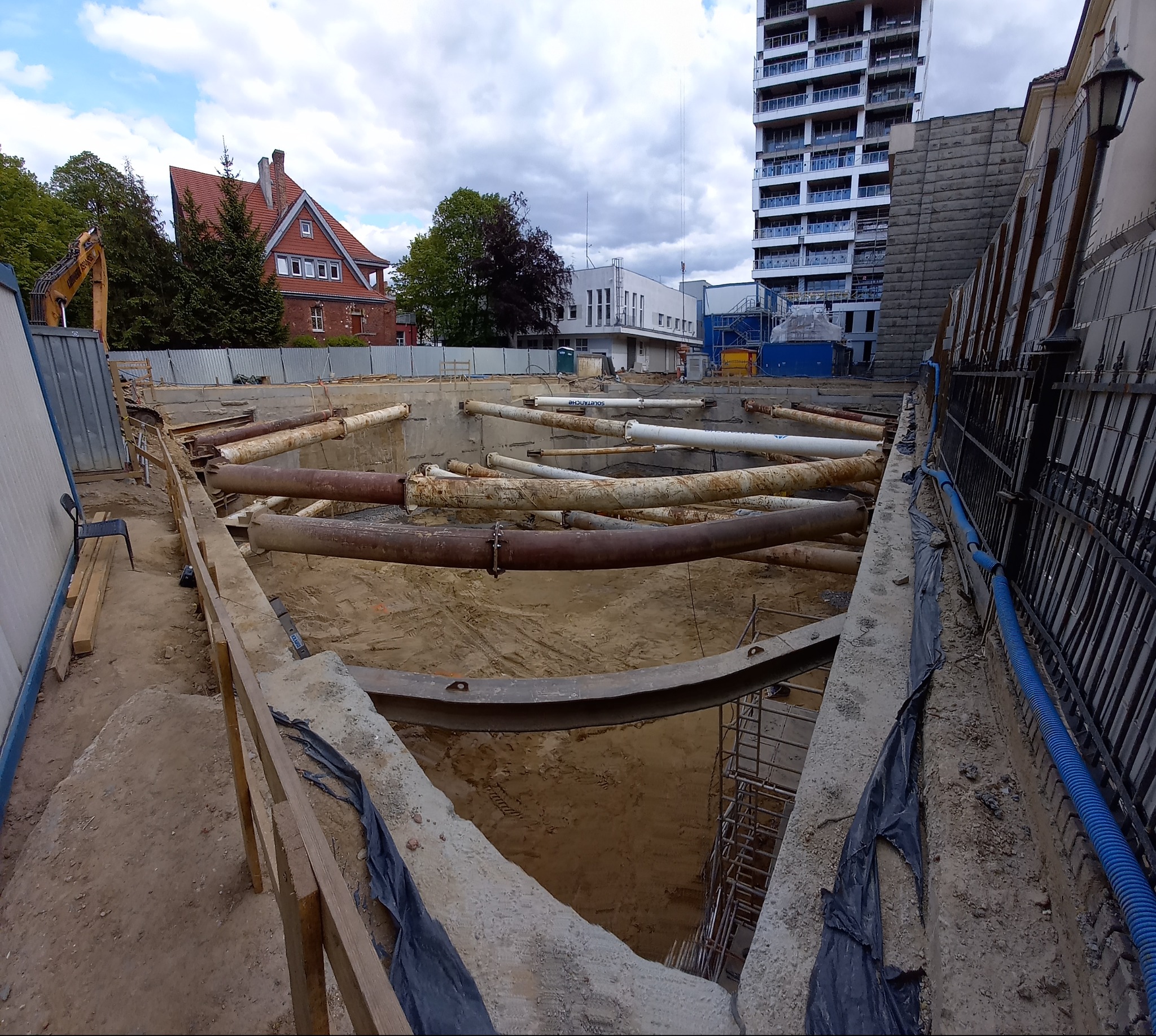 Wykonanie wykopu pod budowę z zabezpieczeniem ścian wykopu rurami stalowymi. W tle budynki mieszkalne i biurowe. Widok z góry na plac budowy.