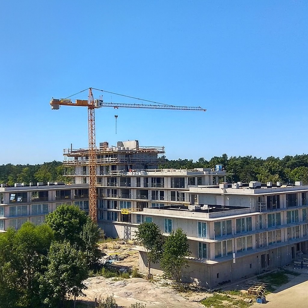 Wysoki budynek w trakcie budowy, widoczny żuraw budowlany na tle błękitnego nieba i zieleni drzew. Budynek z licznymi oknami i balkonami w surowym stanie.