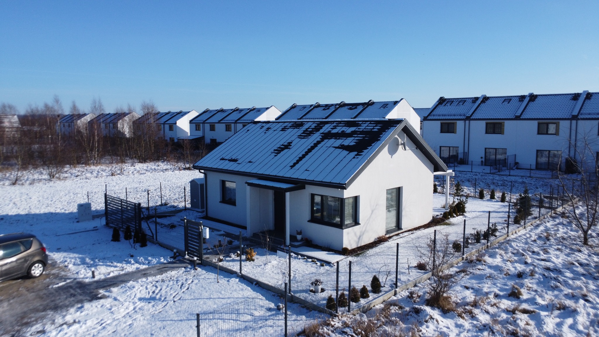 Nowoczesny, biały dom jednorodzinny z ciemną dachówką, otoczony śnieżnym krajobrazem i ogrodzeniem. Widok z góry, w tle szereg identycznych domów.