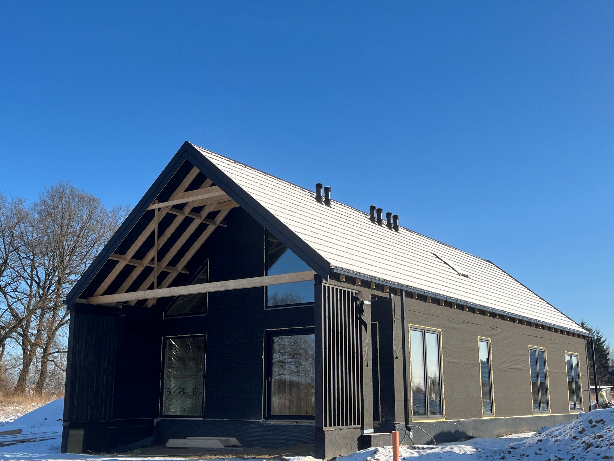 Dom w trakcie budowy, z widoczną konstrukcją dachu i oknami. Elewacja pokryta czarną membraną, dach częściowo ośnieżony. Błękitne niebo w tle.