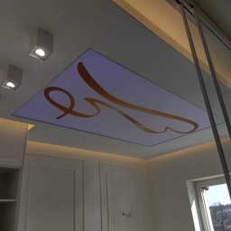 Usługi Remontowo Budowlane Kamil Krawczyk - Nowoczesny sufit podwieszany z podświetleniem LED i abstrakcyjnym złotym wzorem, widok z dołu w jasnym pomieszczeniu z białymi meblami i dużym oknem.