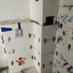 Usługi Remontowo Budowlane Kamil Krawczyk - Montaż białych i wzorzystych płytek w narożniku łazienki, z użyciem klipsów poziomujących i taśmy maskującej, widoczne elementy instalacji wodnej i fragmenty podłoża.
